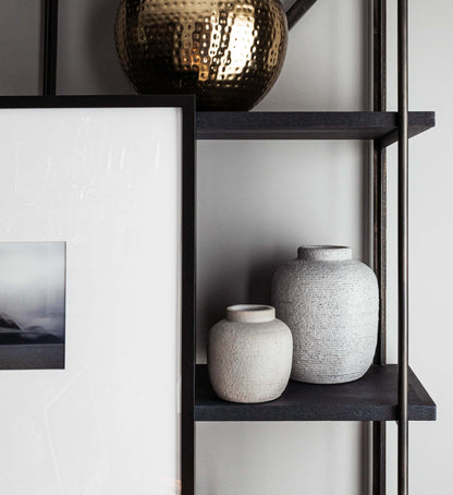 Concrete ribbed and textured vessels/jars/vases on bookshelf with decorative objects.